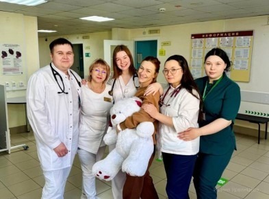 Команда врачей Сургутского кардиоцентра в белых халатах вместе с молодой мамой и сотрудниками клиники, на заднем плане аптечные и информационные стенды.