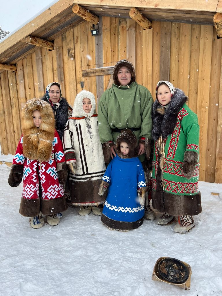 Семья в традиционных нарядах стоит перед деревянным стойбищем: мужчина в зелёном халате, женщины в меховых одеждах, дети в ярких костюмах на фоне снегa