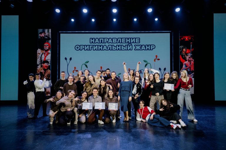 Группа молодых участников на сцене фестиваля Студенческая весна в Сургуте, фон украшен баннером с надписью Направление оригинальный жанр и цветовым оформлениями, отражающим энергичную атмосферу события, на переднем плане улыбающиеся лица